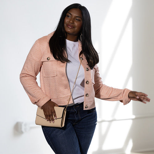 A fashionable woman with long dark hair poses in a peach-colored tweed jacket, white top, and dark denim jeans, accessorized with a neutral crossbody purse. She smiles while looking off to the side in a softly lit room.